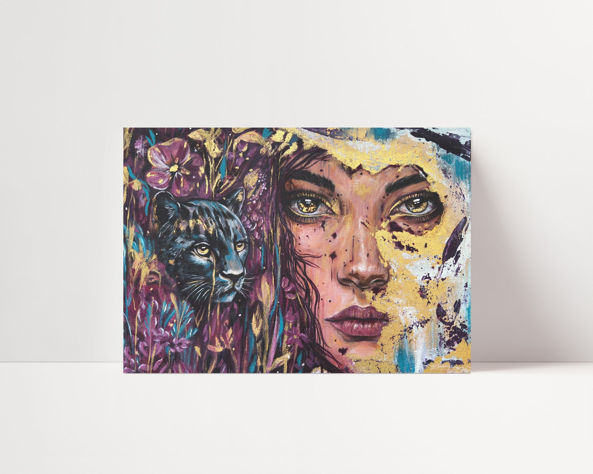 A colorful square  art print leaning against a white wall on a white surface. The artwork depicts a stylized woman's face with a black panther on the left, surrounded by purple flowers and blue, yellow, and pink painterly textures.