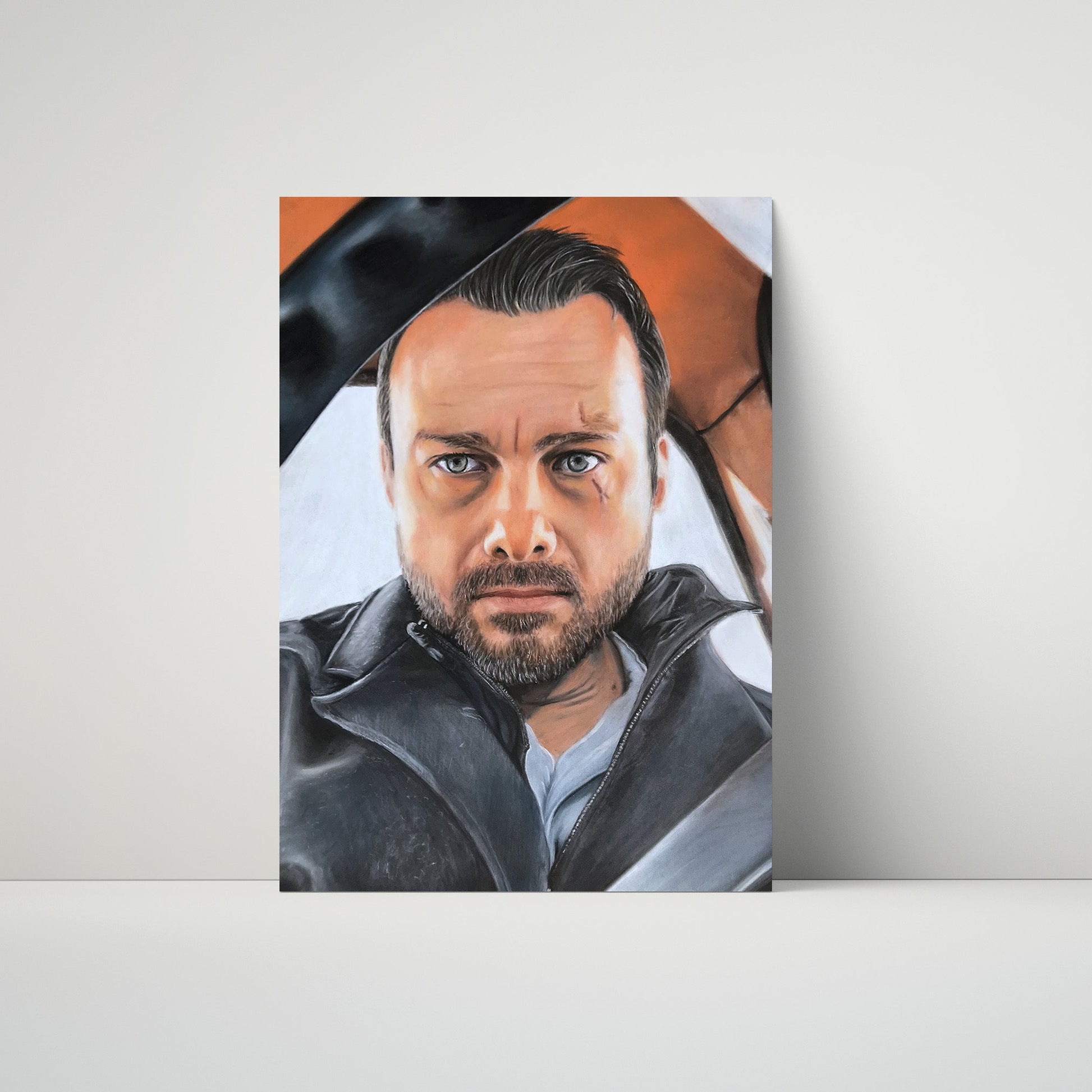 A vertical portrait painting of a man with dark hair, a beard, and blue eyes, wearing a dark jacket, displayed leaning against a light gray wall on a white surface.